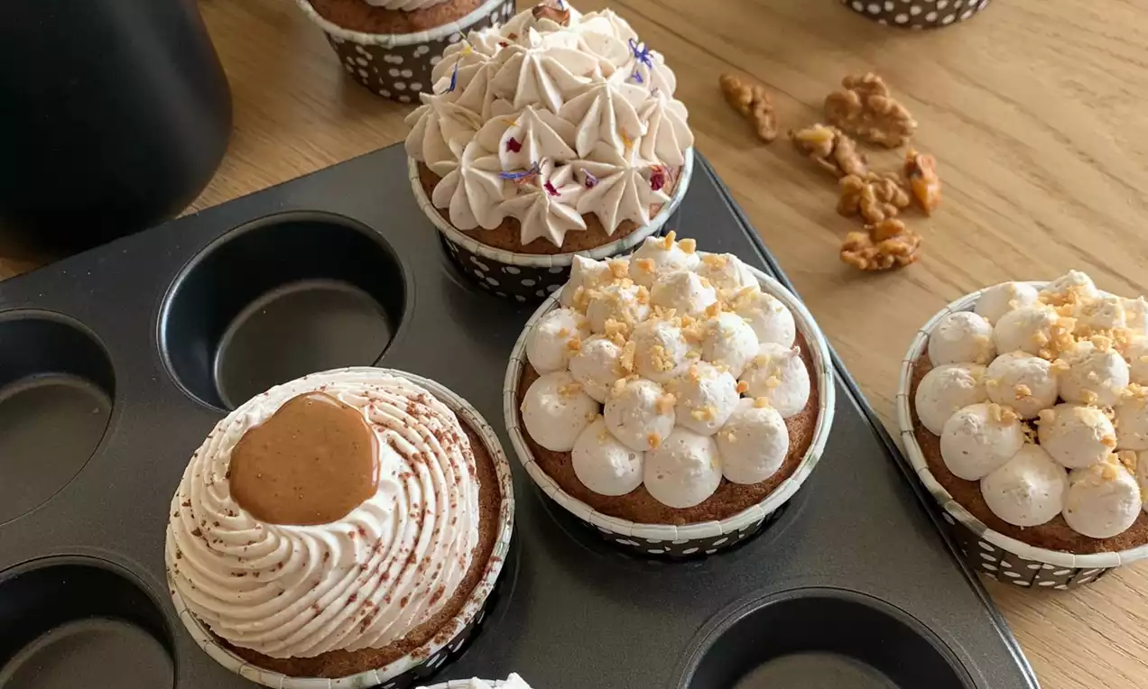 Jednotlivé hnědé muffiny v černobílých papírových košíčcích, zdobené krémovými kopci, ořechy, sušenkou a jedlými květy na plechu