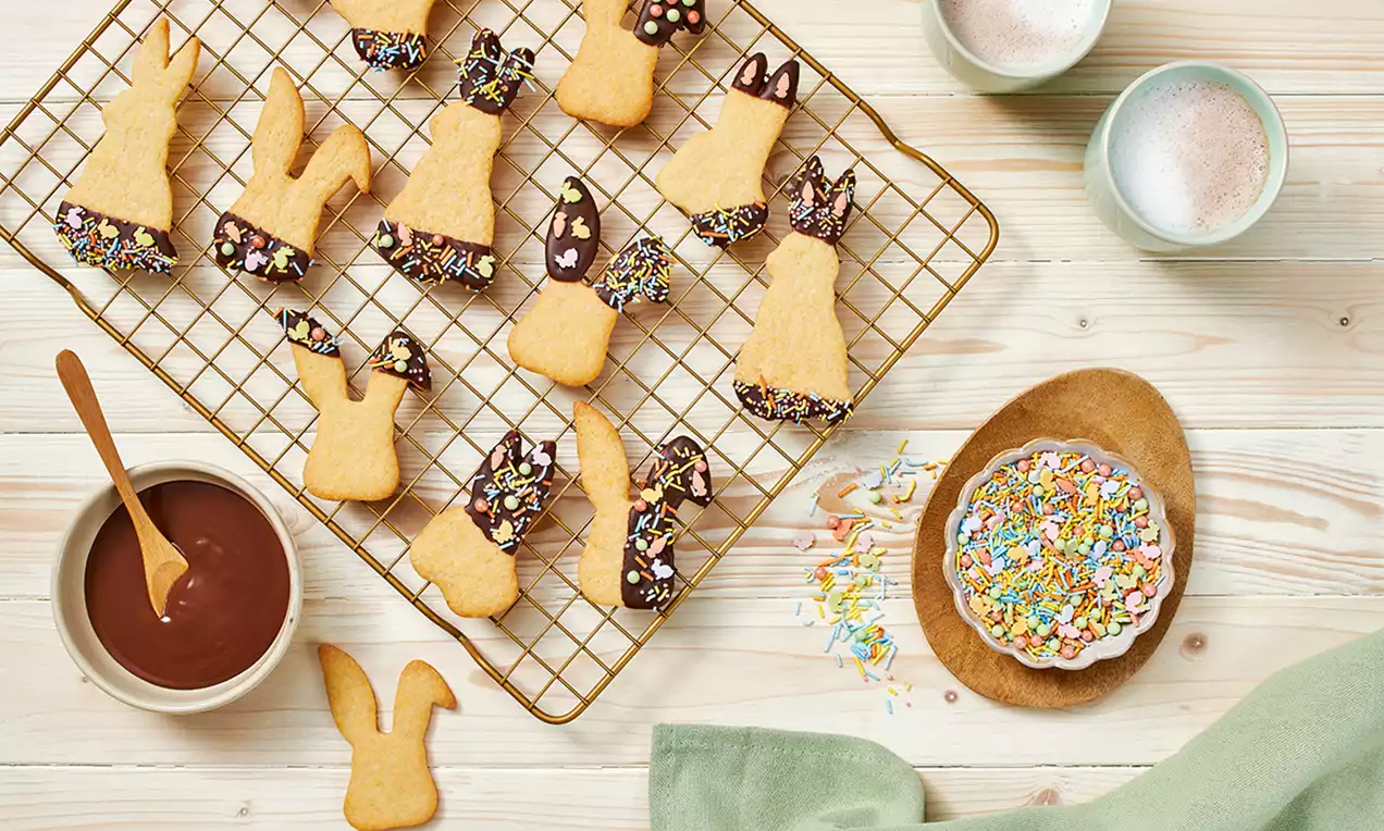 Biscuiți aurii în formă de iepurași, cu urechi și picioare glazurate cu ciocolată, presărate cu ornamente colorate și așezați pe un grătar metalic