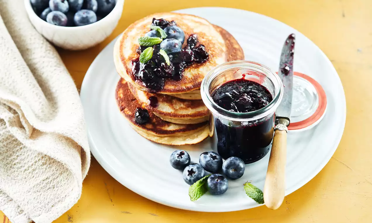 Blauwe bessenconfituur in een glazen pot naast goudbruine pannenkoeken met confituur, verse blauwe bessen en muntblaadjes op een wit bord