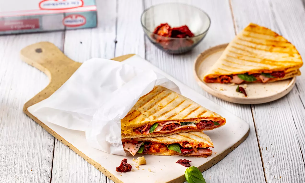 Златисто-кафява триъгълна грил пица с видими слоеве сух домат, зелен лист и тънки резени месо върху бяла хартия на дървена дъска