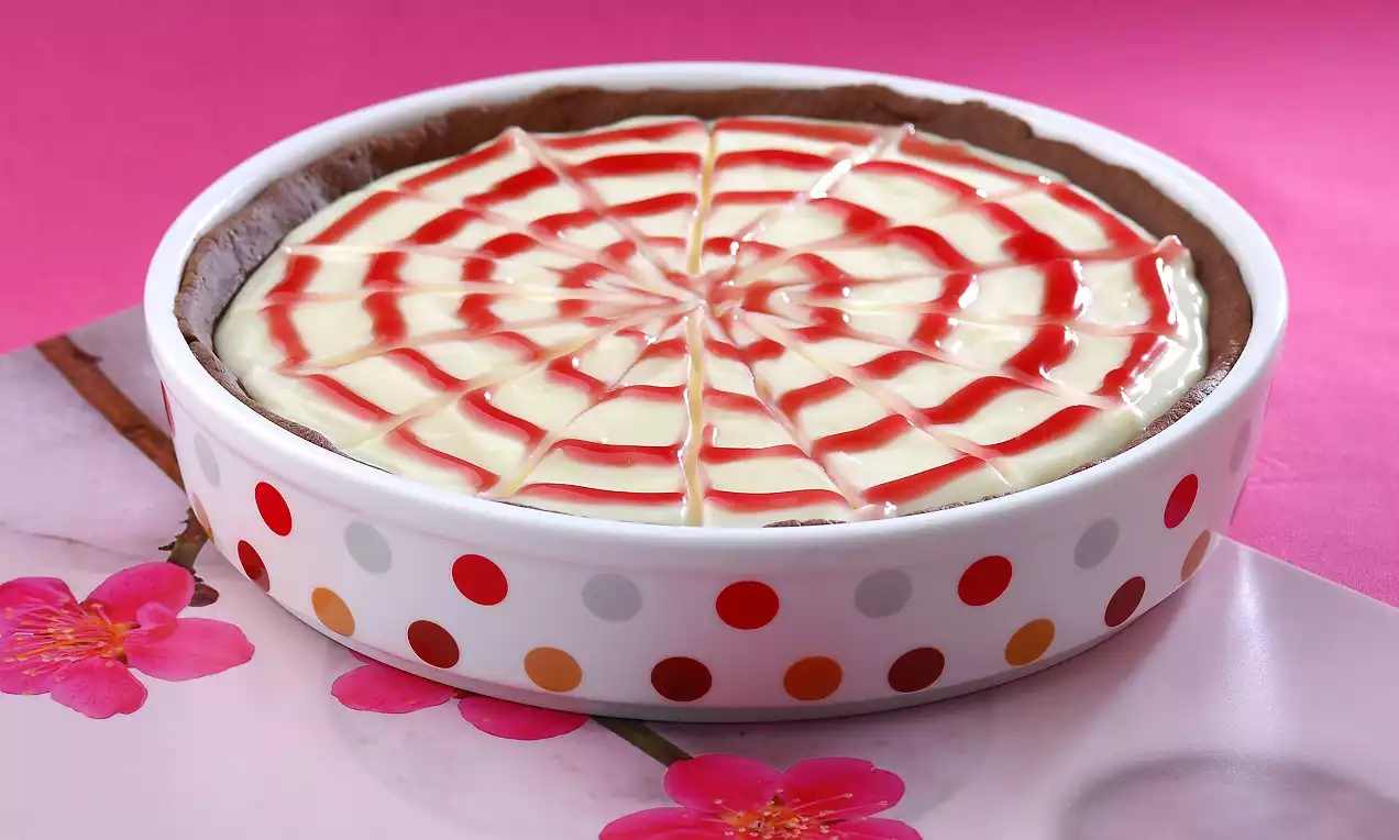Beyaz kırmızı puantiyeli yuvarlak kapta, çilek soslu örümcek ağı deseniyle süslenmiş pürüzsüz beyaz kremalı tatlı. 