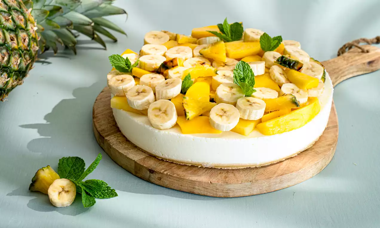 Kwarktaart met een gladde witte vulling, belegd met gele mango-, bananenschijfjes en muntblaadjes, gepresenteerd op een houten plank