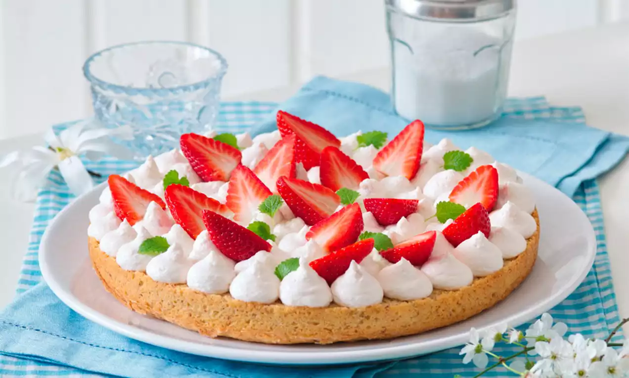 Tvarohový koláč s dozlatova upečeným základem, vrstvou bílého krému, červenými jahodami a lístky máty na bílém talíři na modrém ubrusu