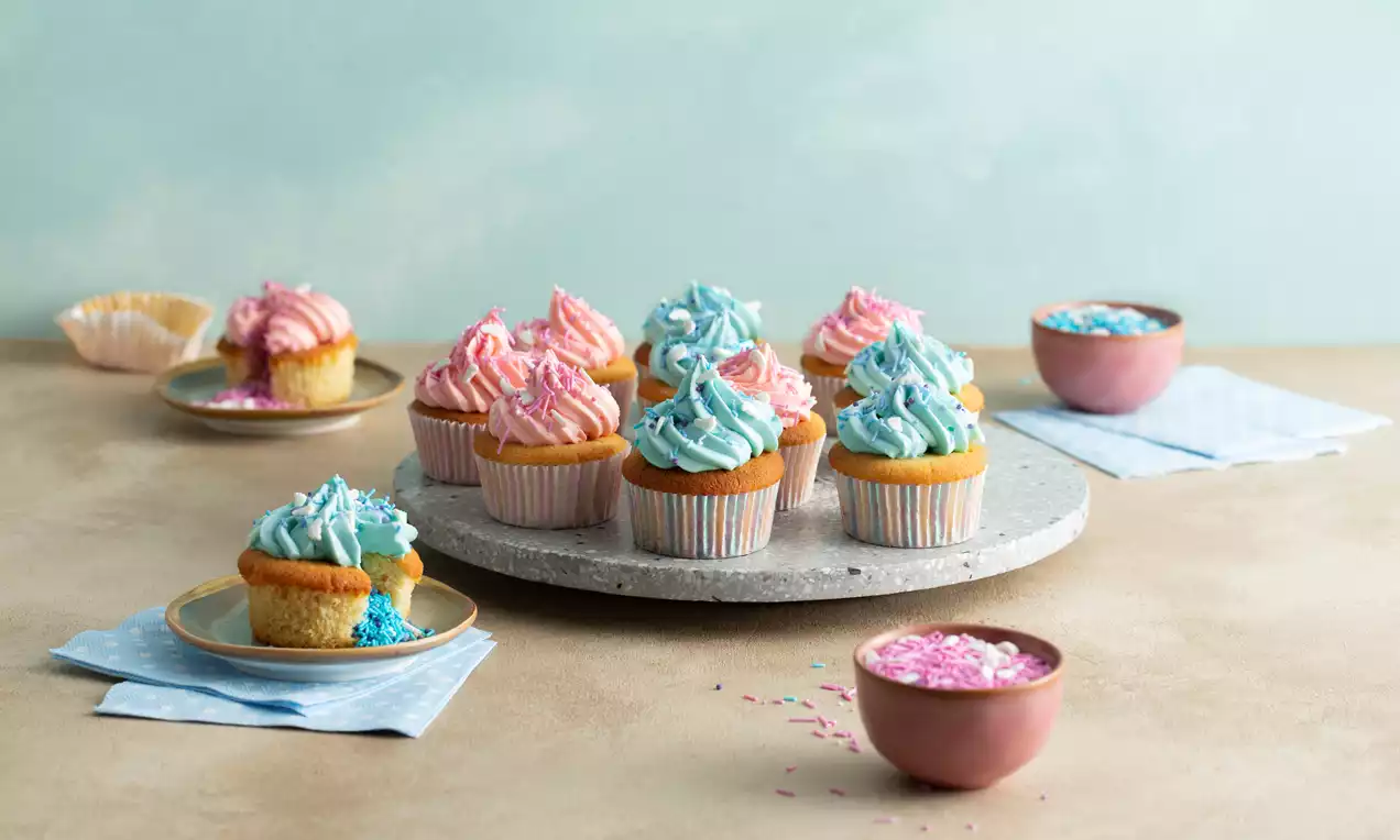Cupcakes met lichtroze en lichtblauwe toefjes botercrème op een grijs dienblad, omringd door schaaltjes strooisel en pastelkleuren servetten