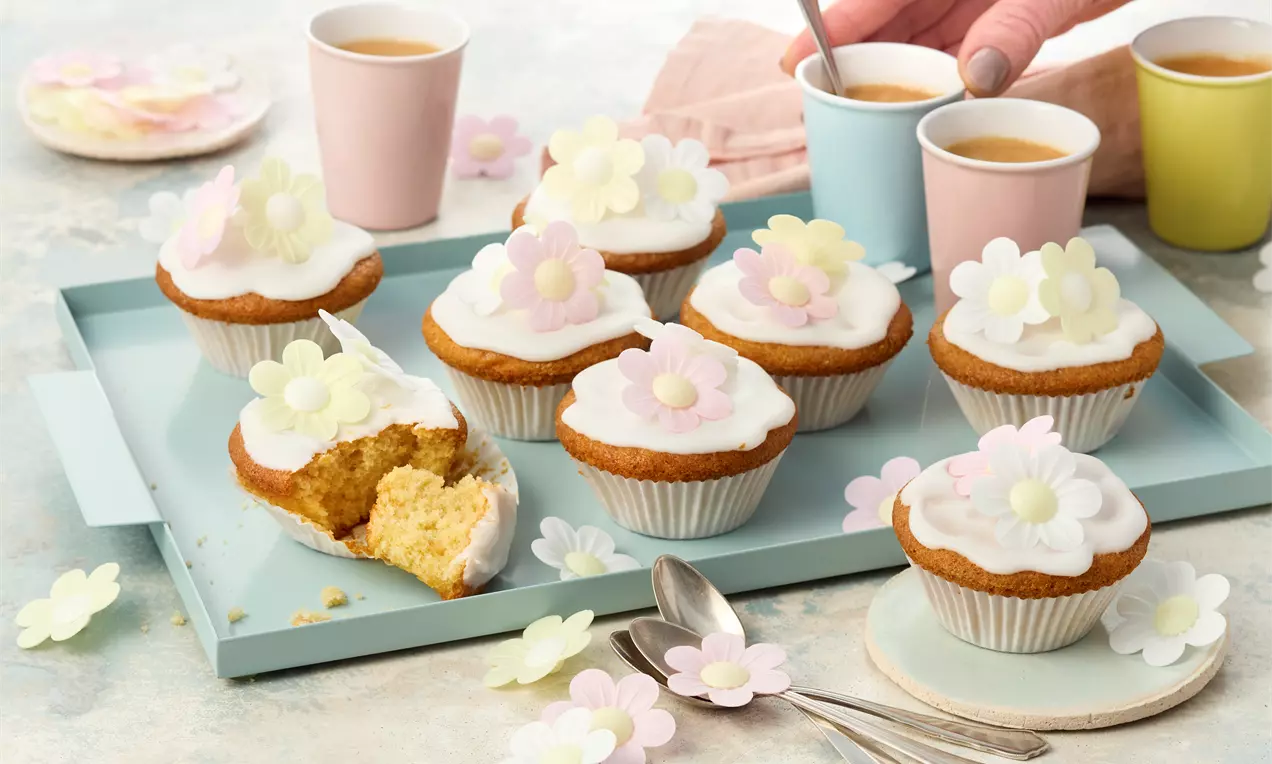 vegane Muffins in weißen Papierbackförmchen, heller Rührteig, weißer Zuckerguss mit pastellfarbenen Esspapierblüten.