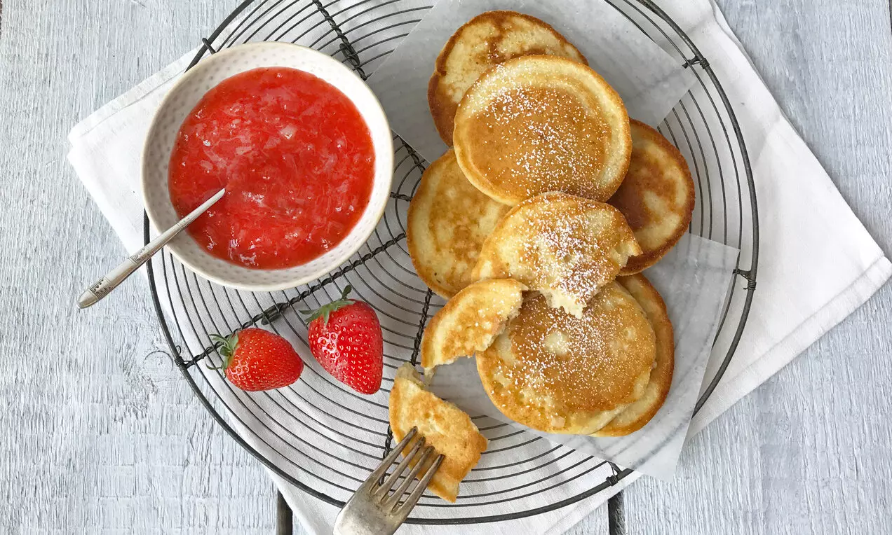 Ярък ягодов конфитюр с ревен в бяла купичка със сребриста лъжица, с гладка текстура, до златисти палачинки върху метална решетка