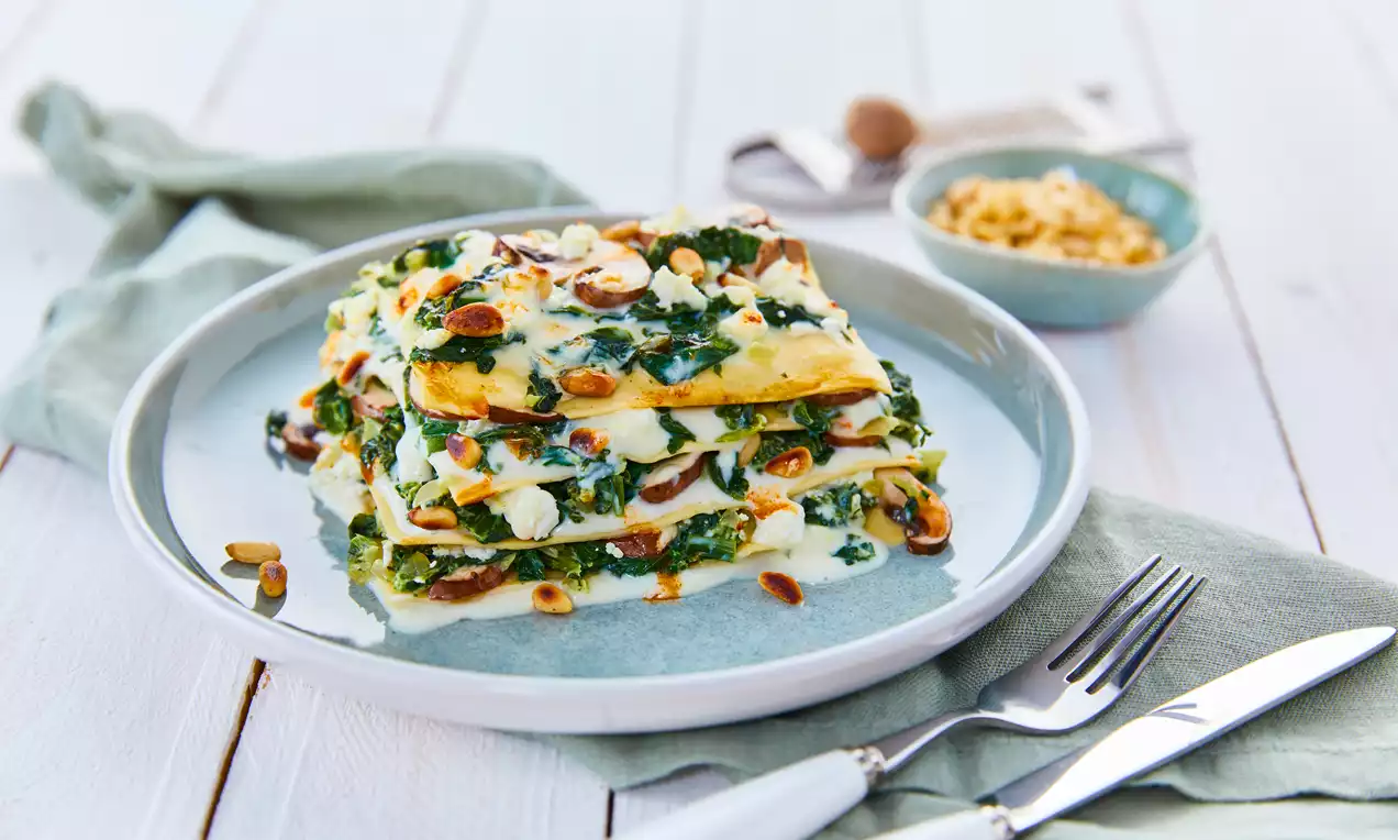 Spinat-Schafkäse-Lasagne mit geschichteten gelben Nudelplatten, grünem Spinat, weißer Sauce und gerösteten Nüssen auf Keramikteller