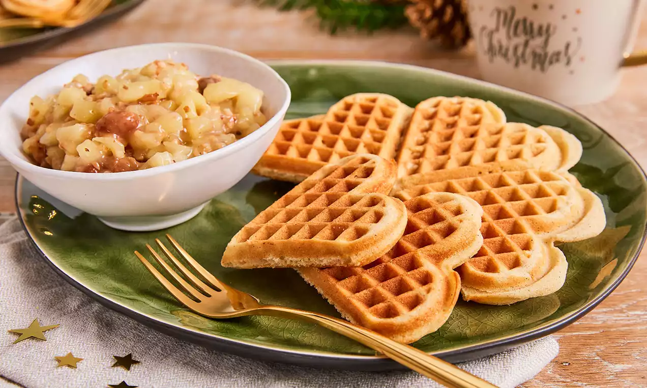 Kalp şeklinde altın rengi waffle'lar, yanlarında tarçınlı elma ve kuru üzümlü bir kaseyle yeşil bir tabakta servis edilmiş
