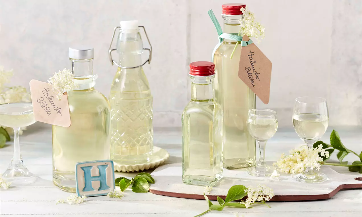 Flaschen mit klarem, hellgoldenem Holunderblütenlikör und geschwungenen Etiketten, dekoriert mit weißen Blüten und grünen Blättern