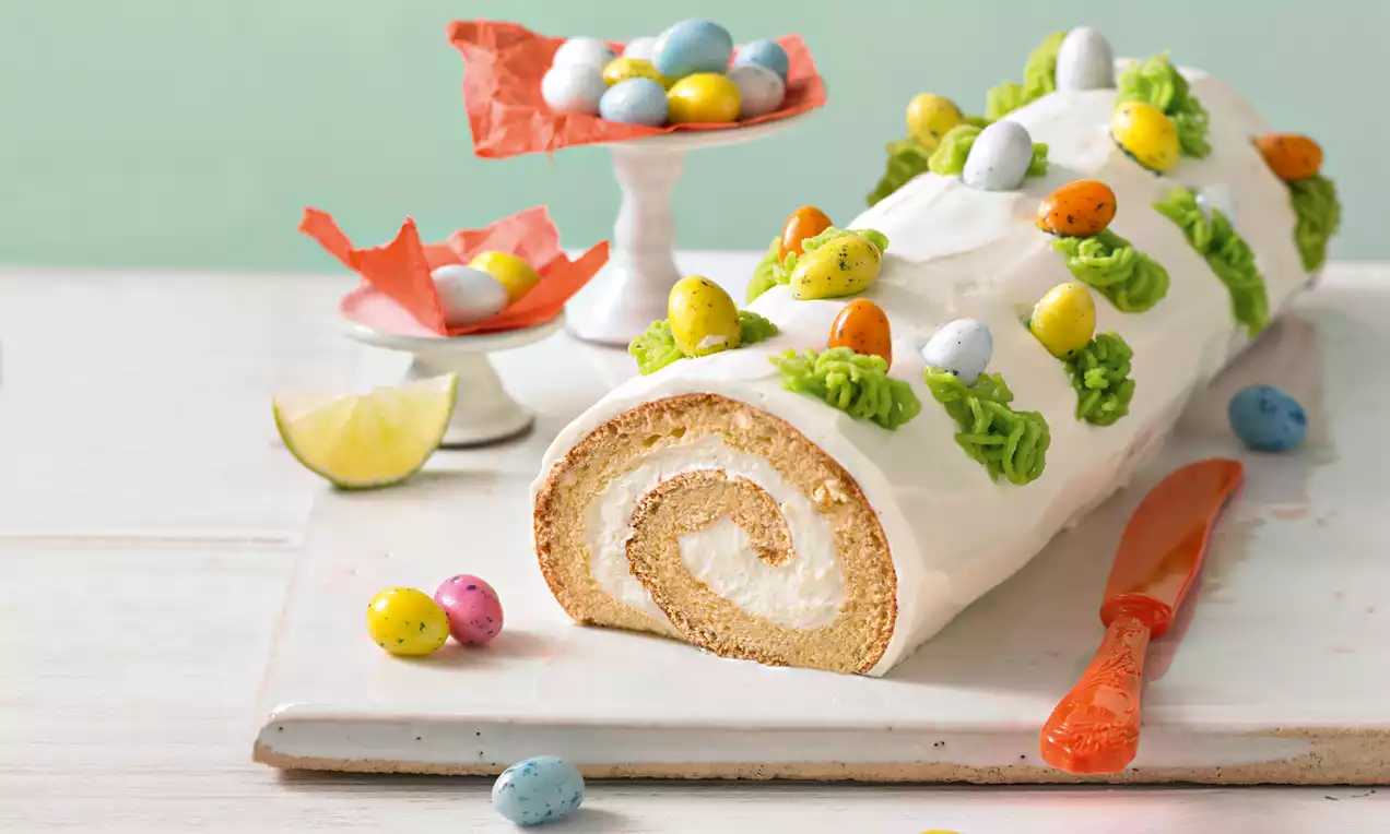 Opgerolde cake met lichte vulling, omhuld met witte crème, versierd met groene toefjes en kleurrijke ei-vormige decoraties op een wit bord
