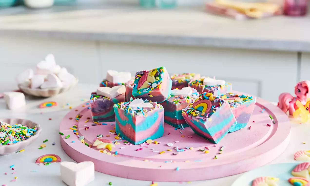 Brightly swirled pink, blue and purple fudge squares topped with rainbow sprinkles on a round pink tray, surrounded by marshmallows