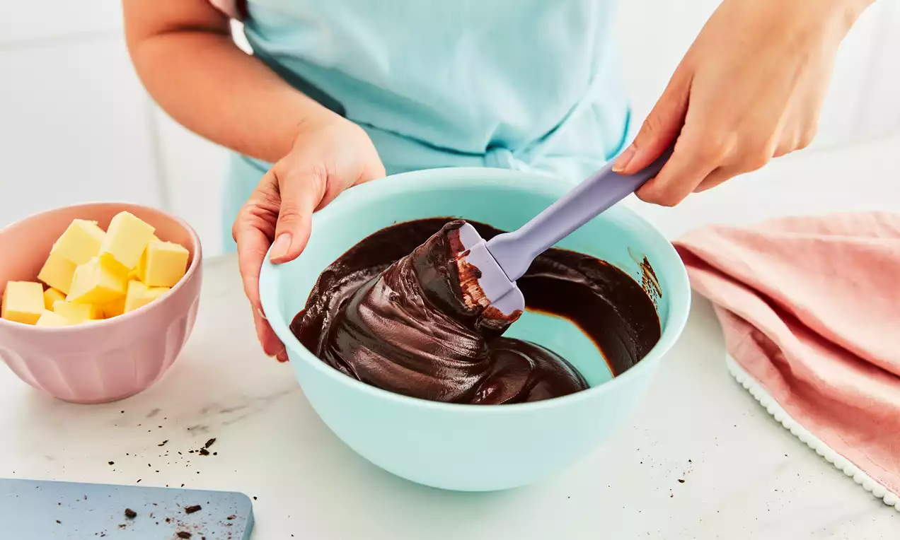 Smooth, glossy dark brown ganache swirled with a lilac spatula in a pale blue bowl next to a pink bowl of cubed butter on a marble counter