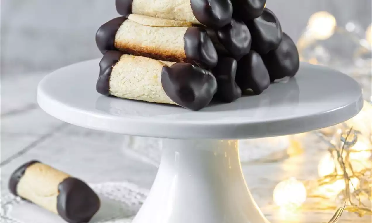 Mandľové trubičky so svetlo zlatou textúrou, oba konce namáčané v tmavej čokoláde, uložené na bielom podnose, obklopené jemnými svetielkami