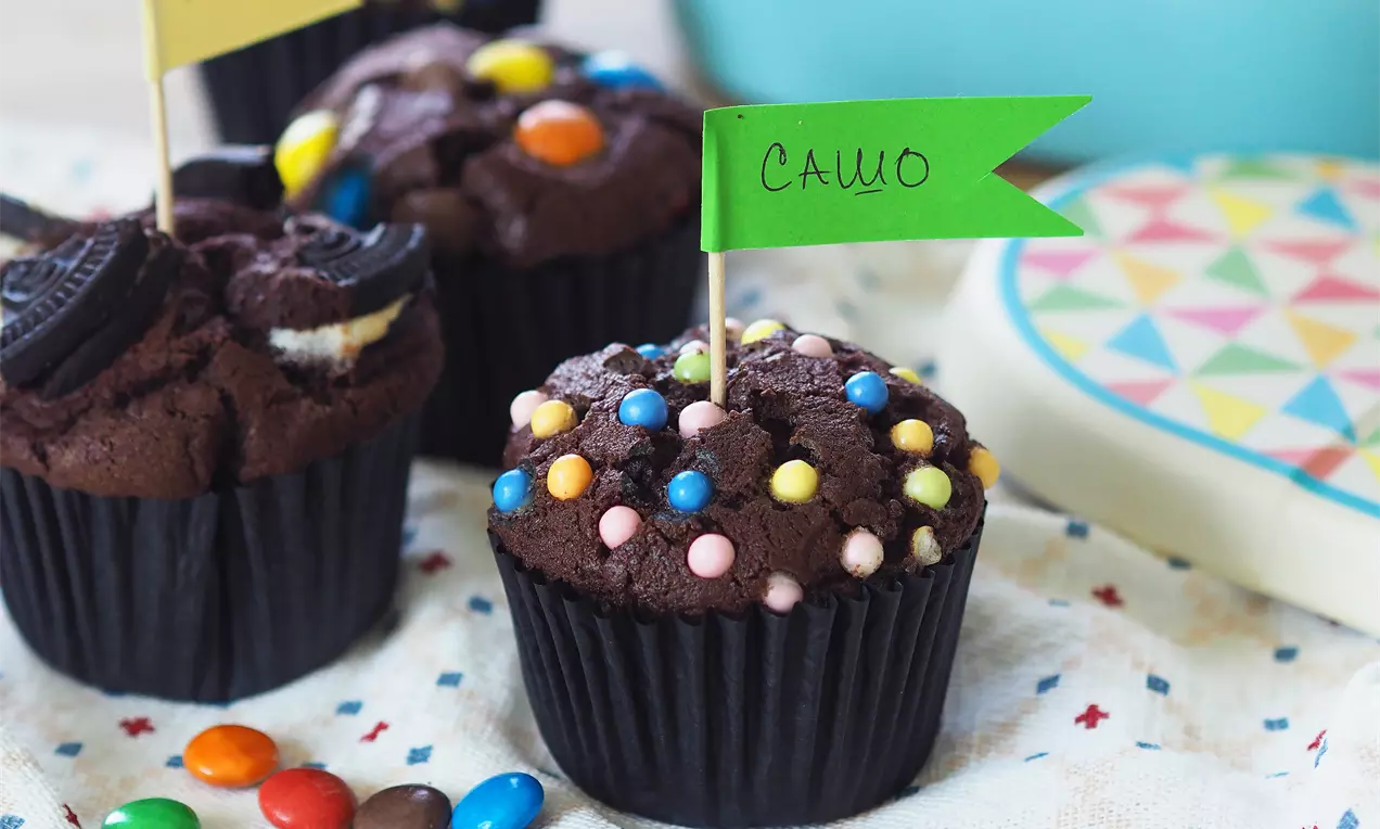 Кафяви шоколадови мъфини с цветни, гладки бонбони на плътна черна хартия, с малки знаменца на пастелна покривка