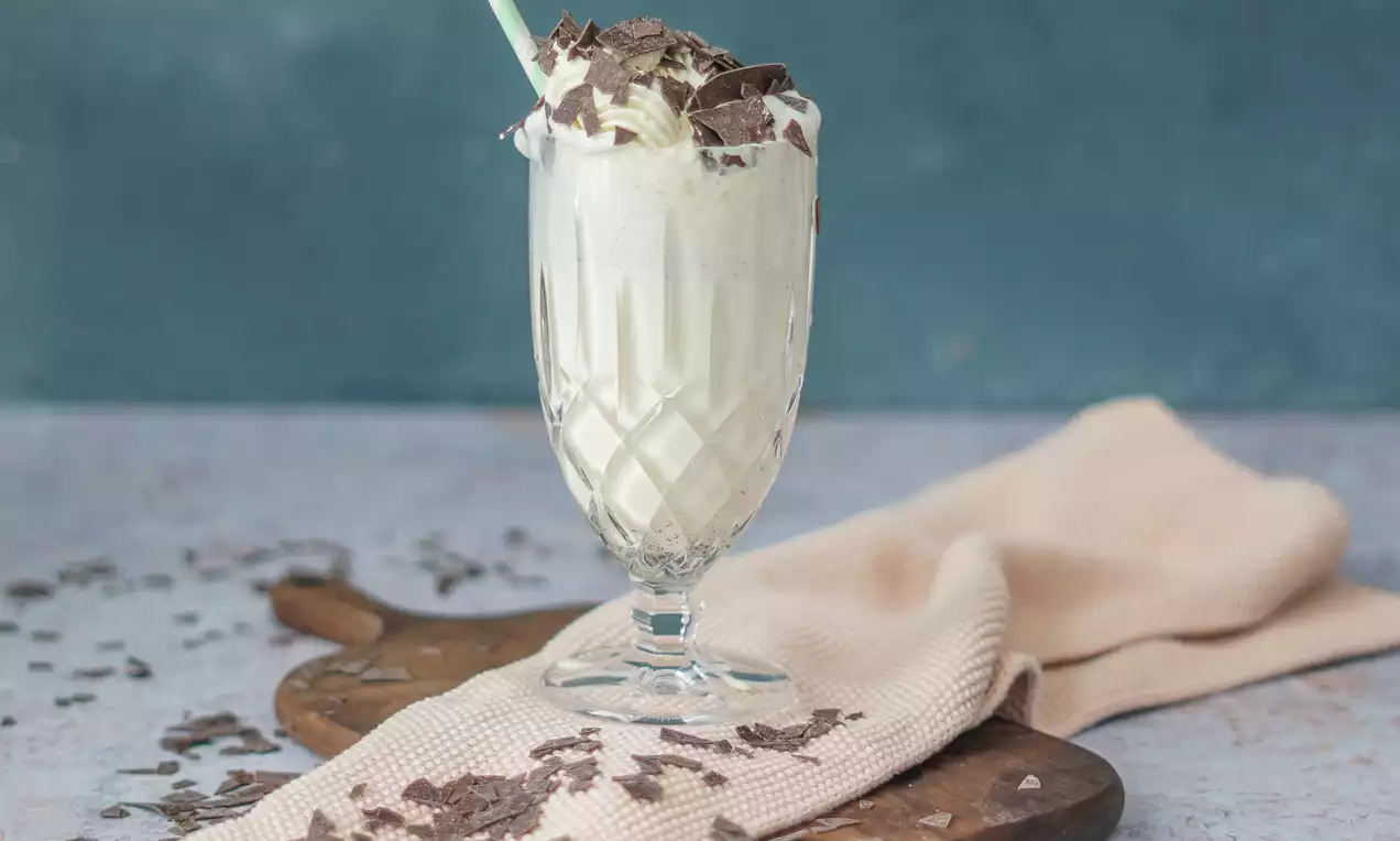 Vanille-Milchshake in einem gläsernen Kelch mit tiefer Rillenstruktur, garniert mit Sahne, Schokoladenraspeln und einem weißen Trinkhalm