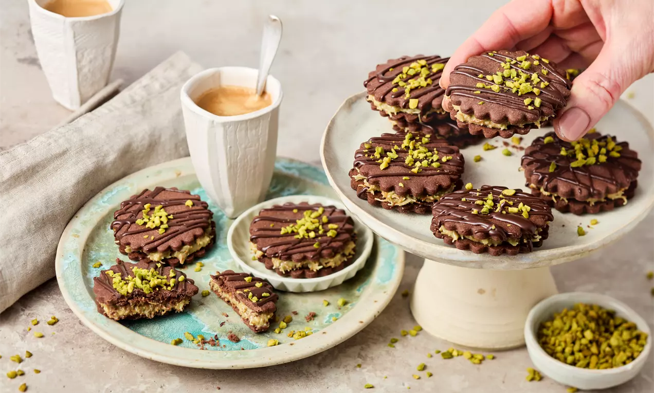 Suklaakuorrutteisia kauniisti uurrettuja keksejä, joita koristavat pistasiamurut, aseteltuna kerrostarjottimelle ja lautaselle cappuccinon kera