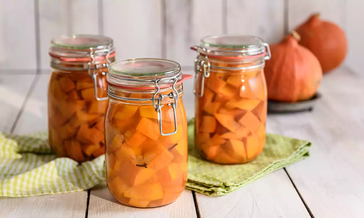 Leuchtend orange Kürbisstücke in klaren Einmachgläsern mit silbernen Bügelverschlüssen, stehend auf einem Holztisch mit grünen Stoffservietten