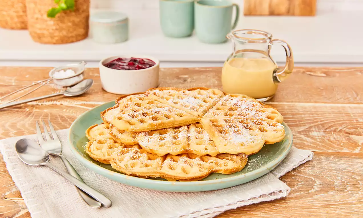 Waffeln ohne Ei auf weißem Teller: goldbraun gestapelt, mit roten Beeren, Blaubeeren, Klecks veganer Joghurt, Minzblatt und Puderzucker