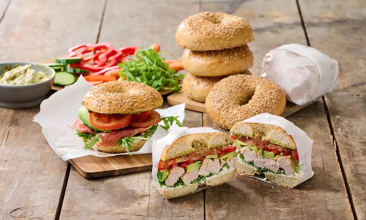 Runde, gyllenbrune sesambagels på en rustikk trebordflate, fylt med sprø salat, avokado, tomat og kylling, med tilbehør av grønnsaker ved siden av