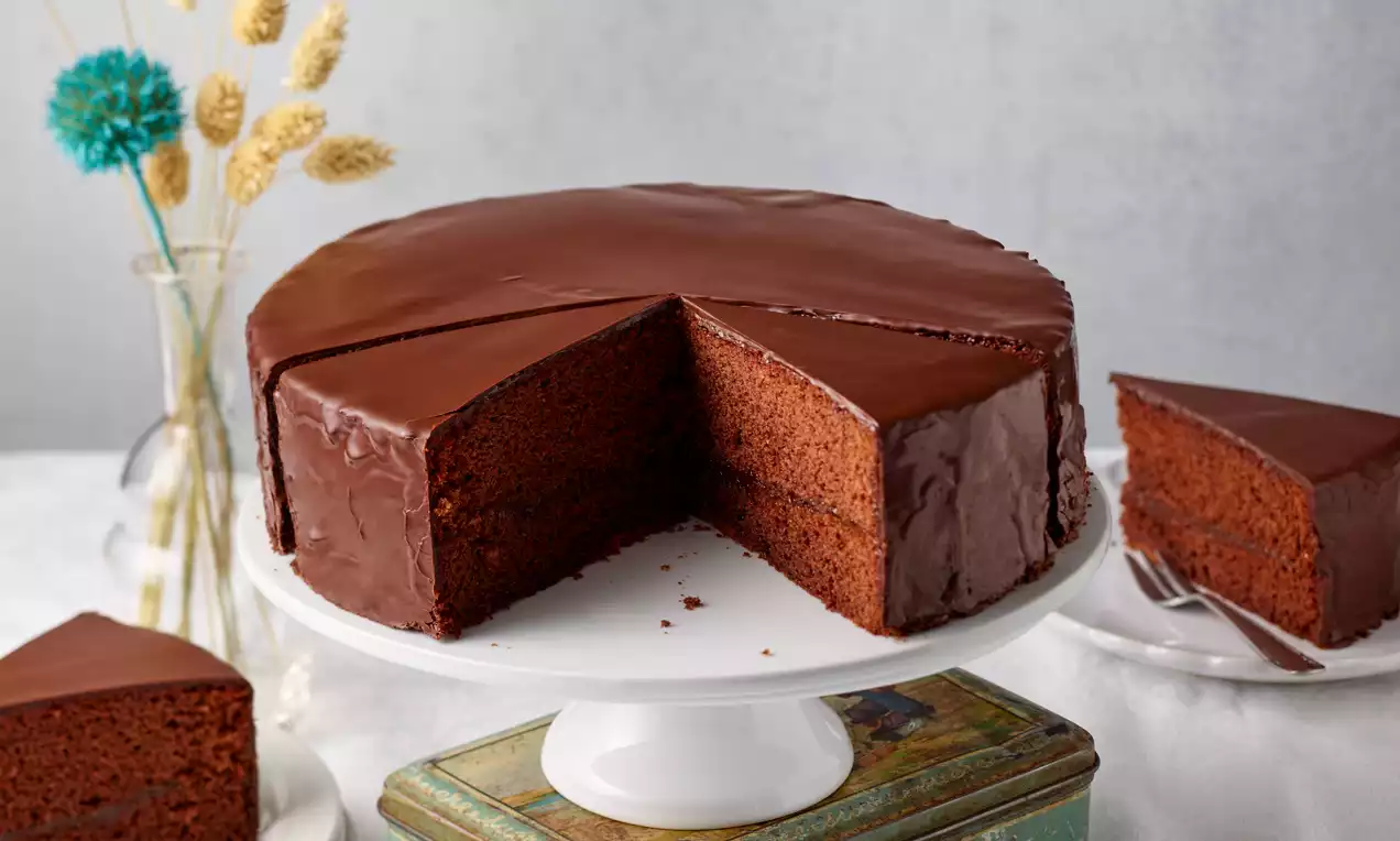 Schokotorte mit glatter, glänzender Glasur und feuchtem, dunklem Teig auf weißer Tortenplatte, daneben angeschnittene Stücke und dezente Dekoration