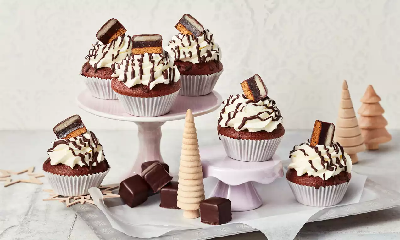 Schokoladige Cupcakes mit weißer Sahnehaube, Schokoladenstreifen und Dominoschwarzstücken auf rosa Etageren mit Dekotannen drapiert