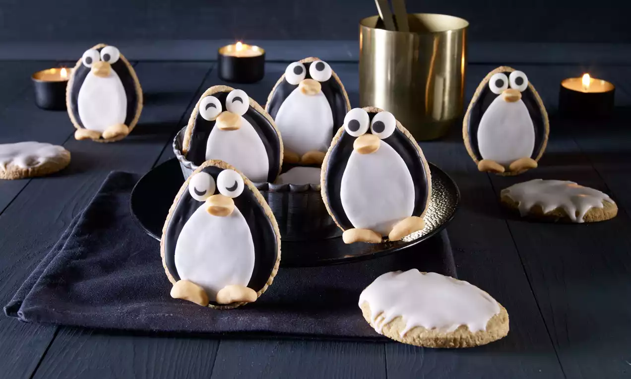 Herzförmige Guetzli mit schwarzem und weißem Zuckerguss wie Pinguine gestaltet, mit großen Augen und orangefarbenen Details auf schwarzem Stofftuch