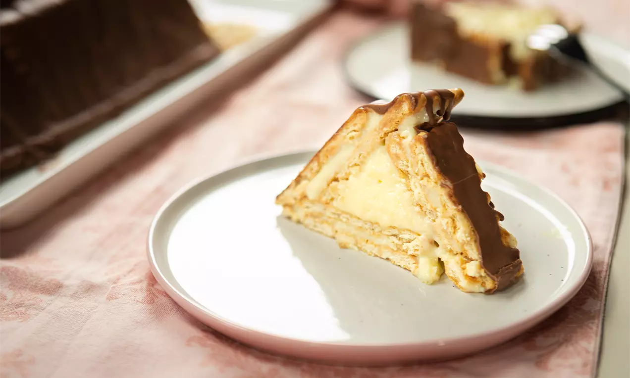 Триъгълно парче с пластове светли бисквити и крем в основа, покрито с гладка кафява глазура върху бяла чиния на розов фон