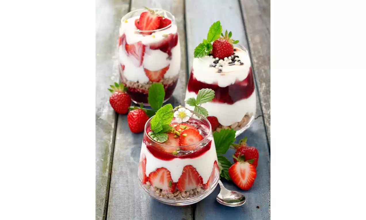 Coupes translucides garnies de couches de crème blanche, fraises rouges entières et tranchées, granola doré et feuilles de menthe verte