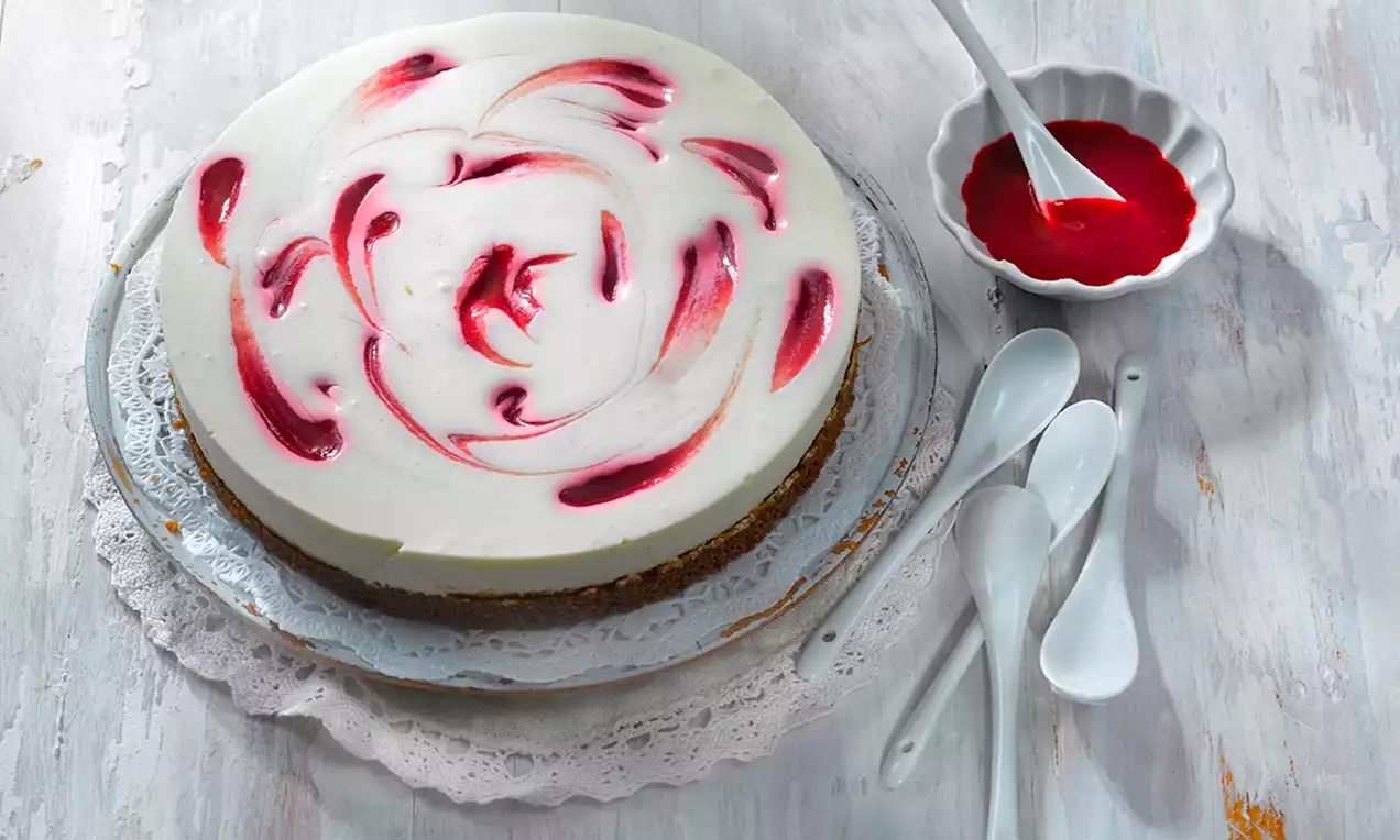 Pyöreä juustokakku vaalealla pinnalla, jossa punaiset pyörteet, aseteltuna pitsikoristeiselle tarjottimelle, lusikoiden vieressä