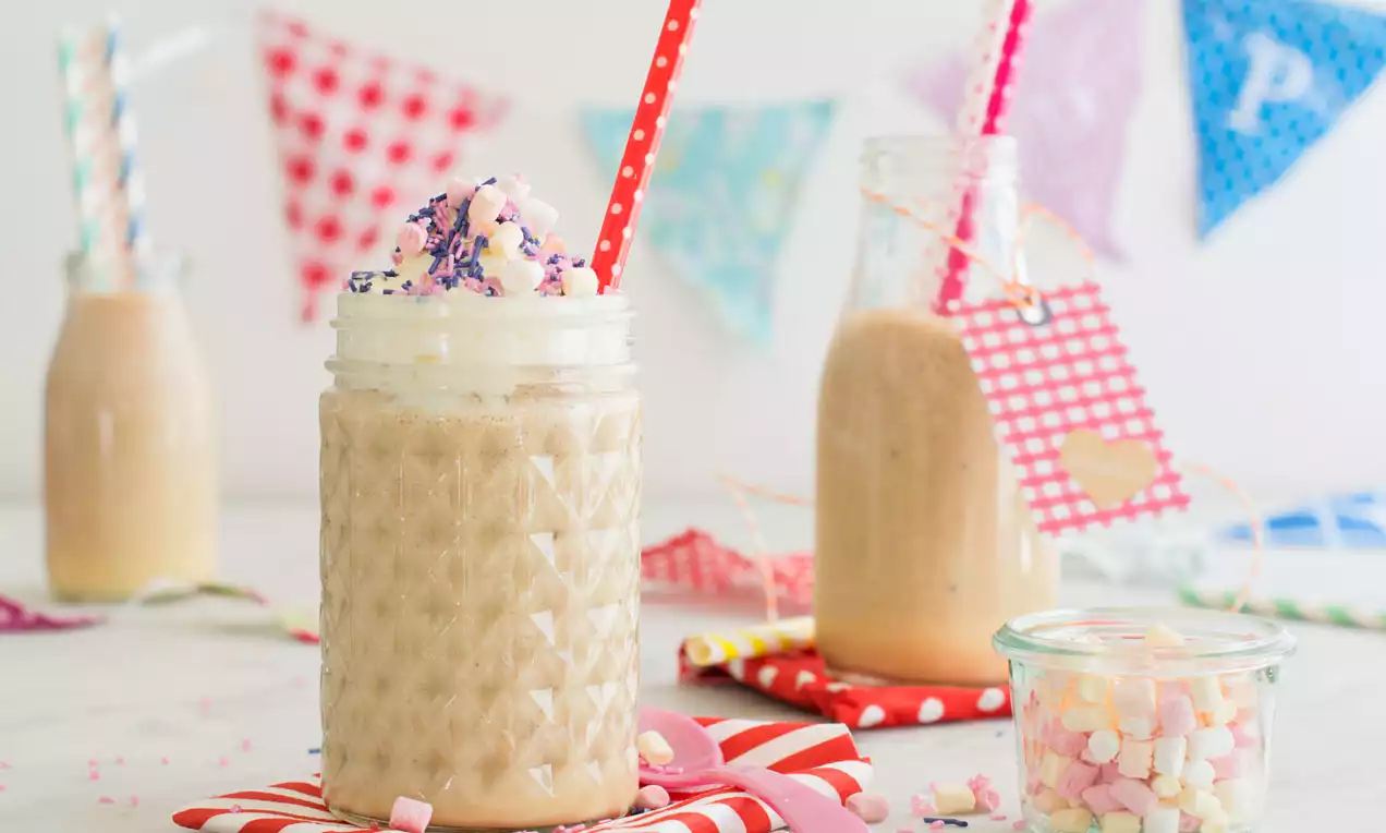 Stracciatella-Shake in einem gläsernen Henkelbecher mit Rautenmuster, gekrönt von Sahne, bunten Marshmallows und Zuckerstreuseln mit rotem Strohhalm