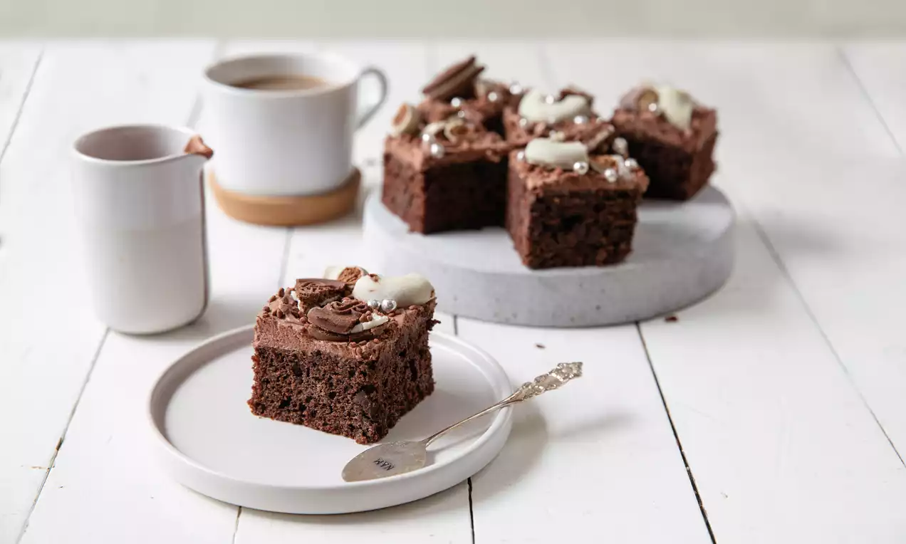 Täyteläisen tummat, pehmeän kosteat neliönmuotoiset suklaaleivokset vaalealla lautasella, koristeena suklaakreemiä ja vaaleita suklaapaloja