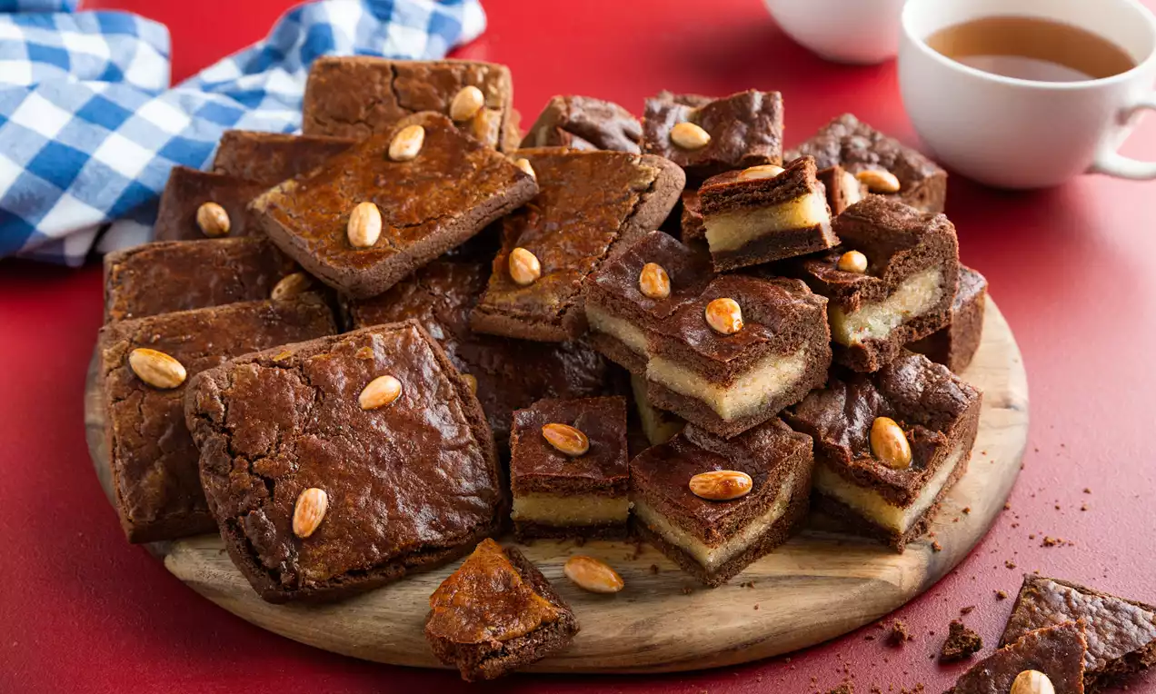 Goudbruine speculaasbrokken met amandelen, rechthoekig en lichtkrokant, gestapeld op een houten bord tegen een rode ondergrond