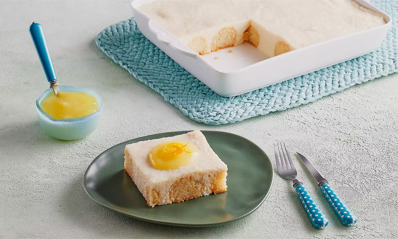 Dikdörtgen dilim irmik tatlısı, üzeri kremamsı beyaz ve ortasında parlak sarı sos, yeşil tabakta, yanında çatal bıçak ile sunulmuş