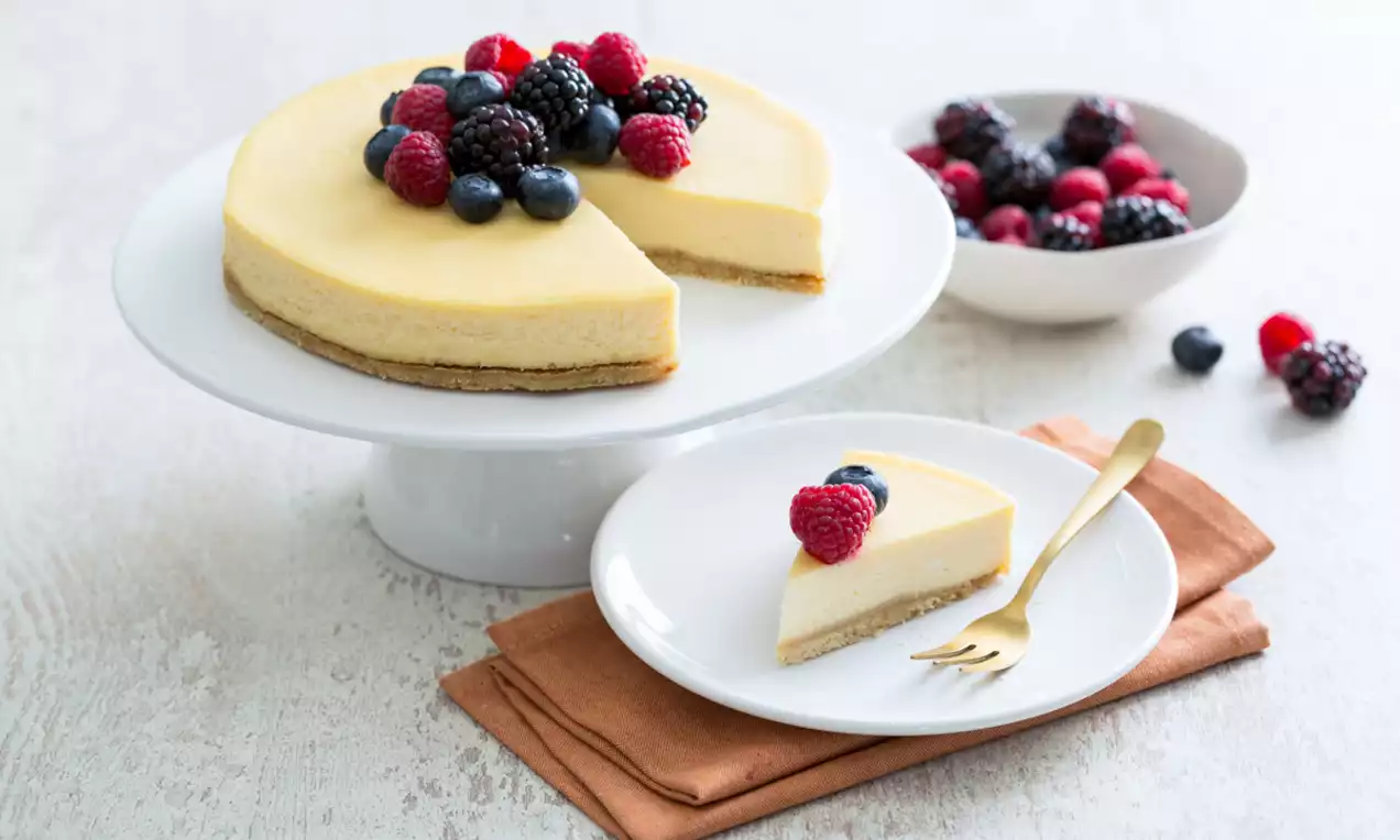 Cheesecake met een gladde, lichtgele bovenkant en een bruine kruimelbodem, gegarneerd met rood en blauw fruit op een witte standaard en bordje