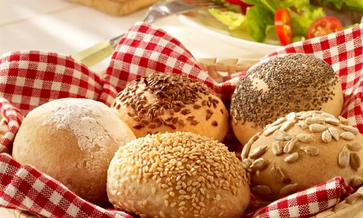 Fünf runde Brötchen mit verschiedenen Samen und Körnern auf rot-weiß kariertem Stoff in einem Korb neben Salat auf weißem Teller