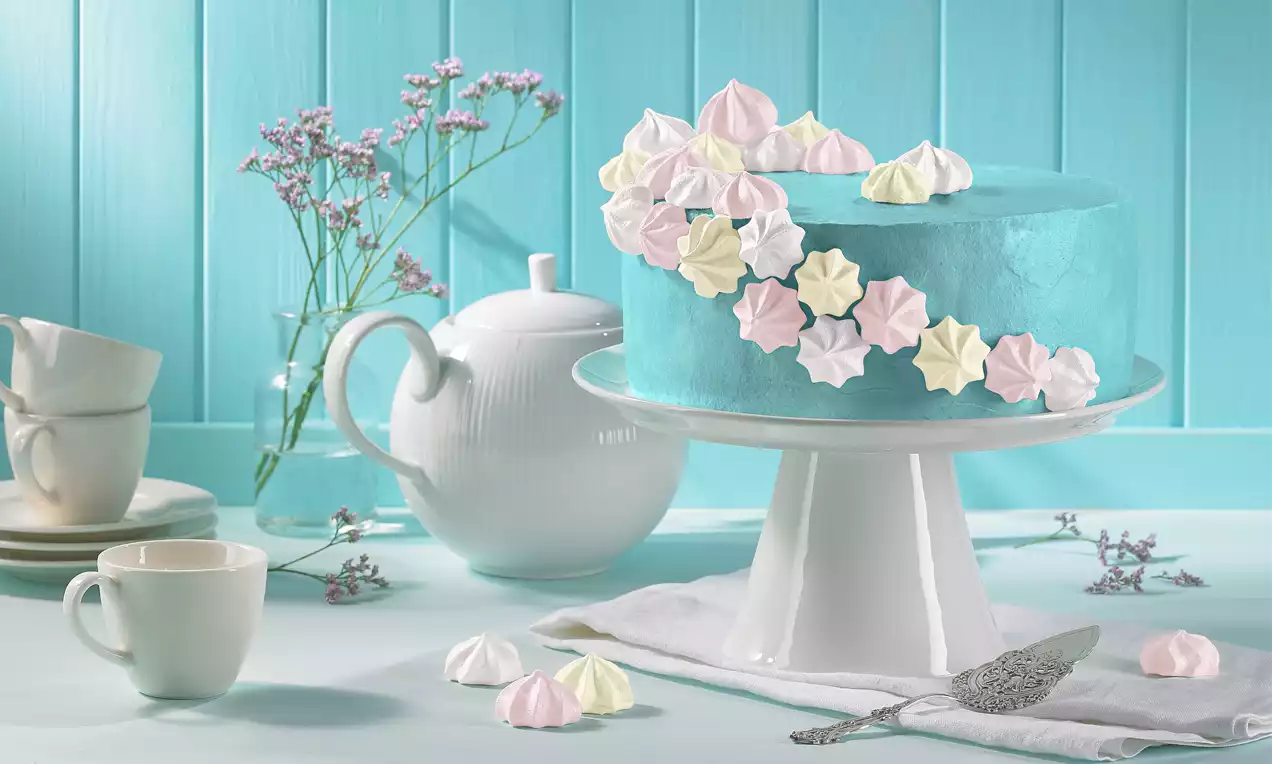 Runde Vanilletorte in hellblauer Creme eingehüllt, mit pastellfarbenen Baisers dekoriert auf weißer Tortenplatte vor blauer Holzoptik