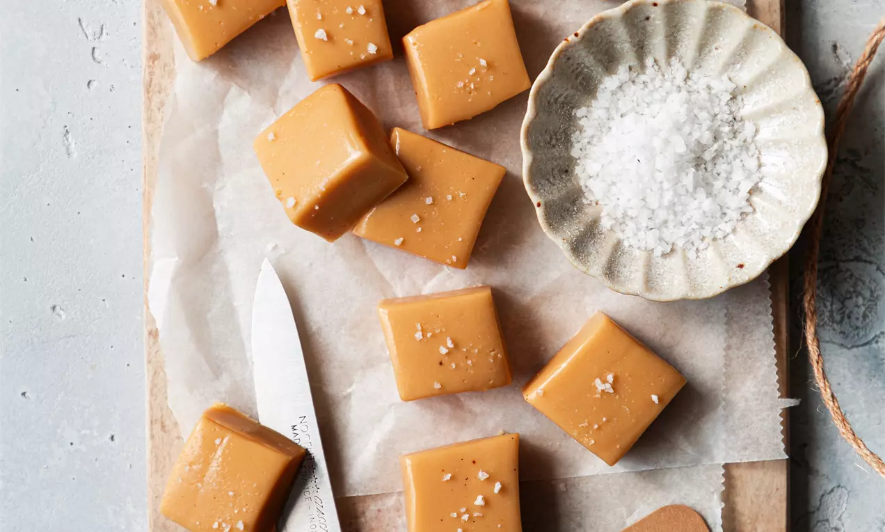 Picture - Golden caramel-coloured vanilla fudge cubes with a smooth surface and sea salt flakes, arranged on parchment with a knife and small dish of salt