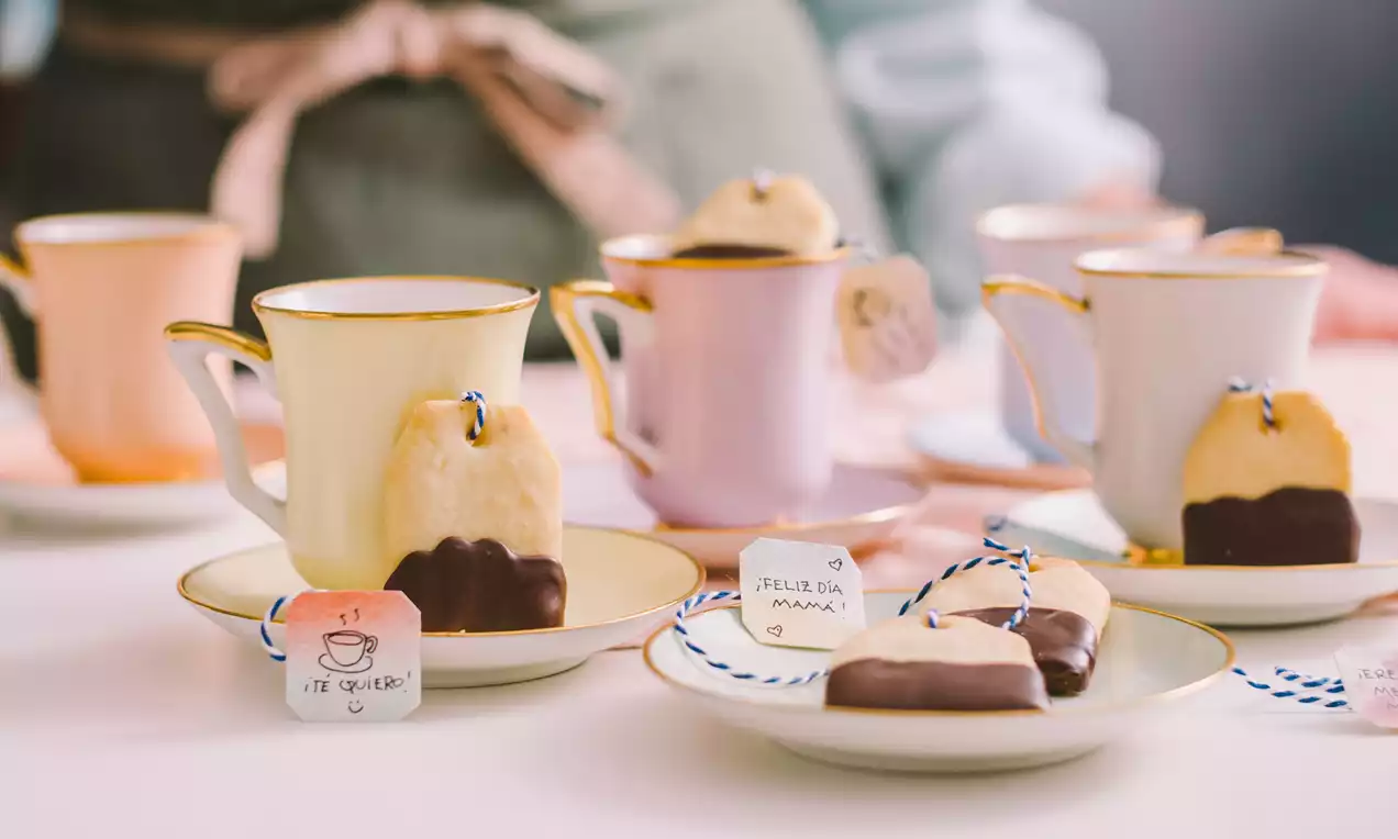 Galletas doradas con puntas bañadas en chocolate oscuro, forma irregular y etiquetas colgadas junto a tazas pastel sobre platos claros