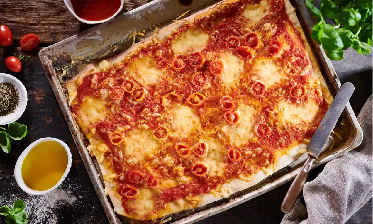 Familienpizza mit rechteckigem Teigboden, intensiv roter Tomatensoße, geschmolzenem Käse und gleichmäßig verteilten Tomatenscheiben in Metallform
