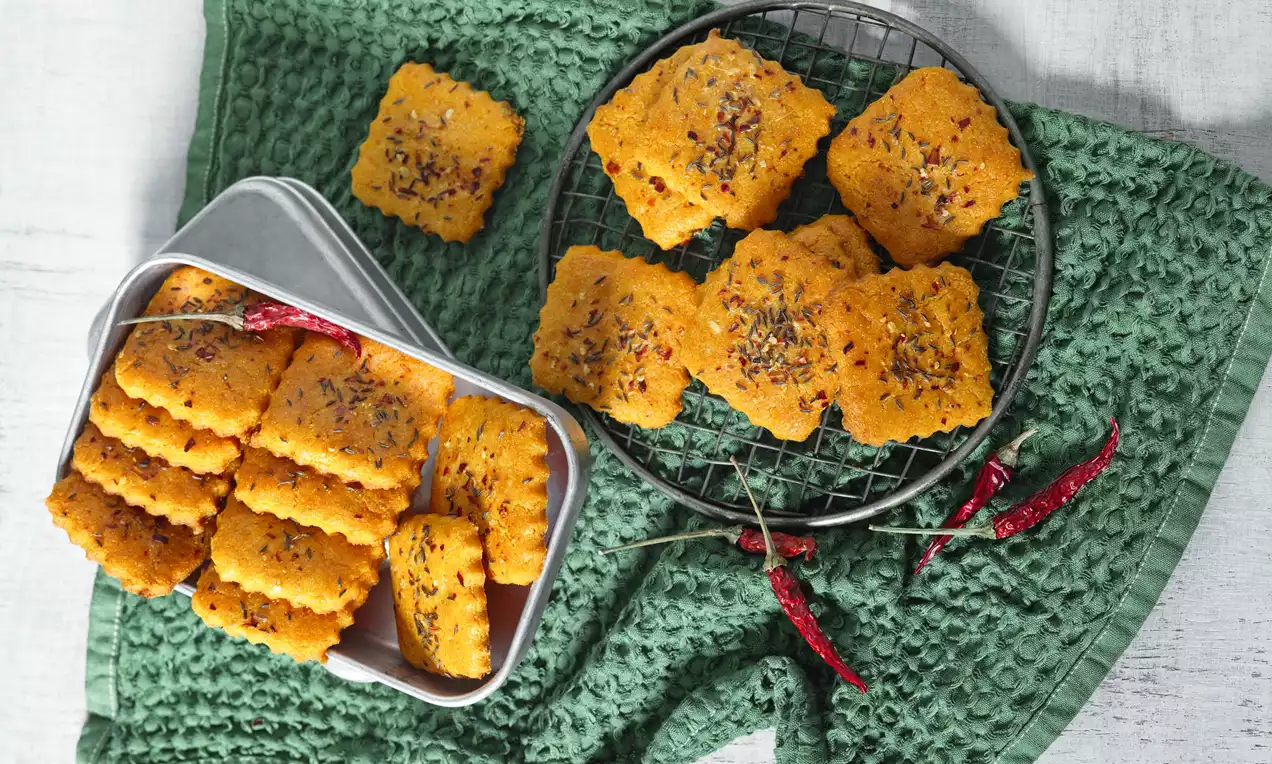 Křupavé čtvercové chlebíčky zlatavé barvy posypané drobnými chilli vločkami, uspořádané na mřížce a v plechové krabičce na zeleném látkovém podkladu