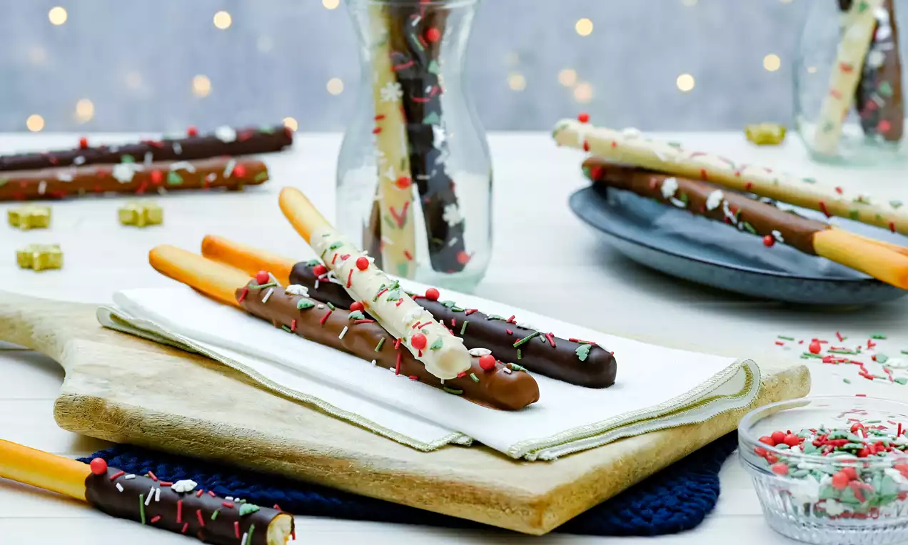 Chokladtäckta sticks i brunt och vitt med färgglada strössel, upplagda på träbräda med servett, glasburk och tallrik i bakgrunden
