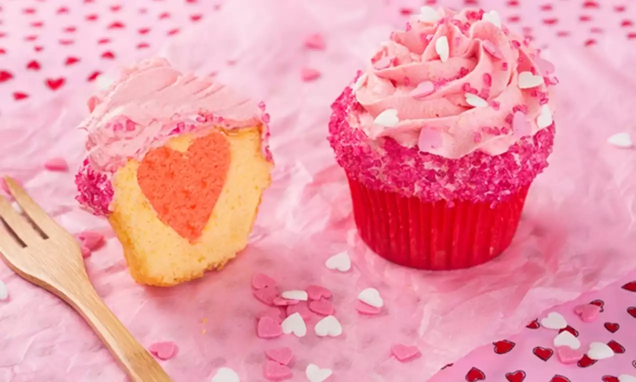 Rosa cupcakes med krämig frosting och hjärtströssel, en delasitu visar ljus sockerkaka med rött hjärta, trägaffel på rosa papper