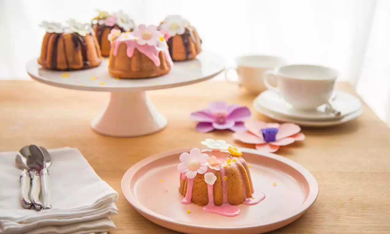 Mini prăjituri aurii, glazurate cu roz și alb, decorate cu flori de zahăr pe platouri elegante albe lângă cești de ceai și tacâmuri argintii