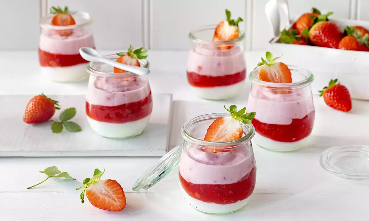 Kleine Gläser mit weißer Skyr-Creme, rotem Fruchtspiegel und rosa Erdbeercreme, garniert mit halbierten Erdbeeren auf weißem Untergrund