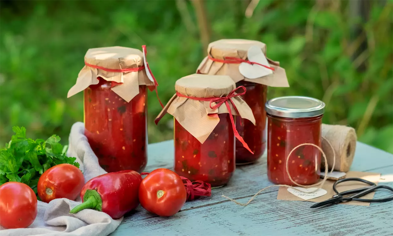 Консервирани домати с наситено червен цвят в стъклени буркани с хартиени капачки и червени панделки, подредени върху дървена маса до пресни зеленчуци