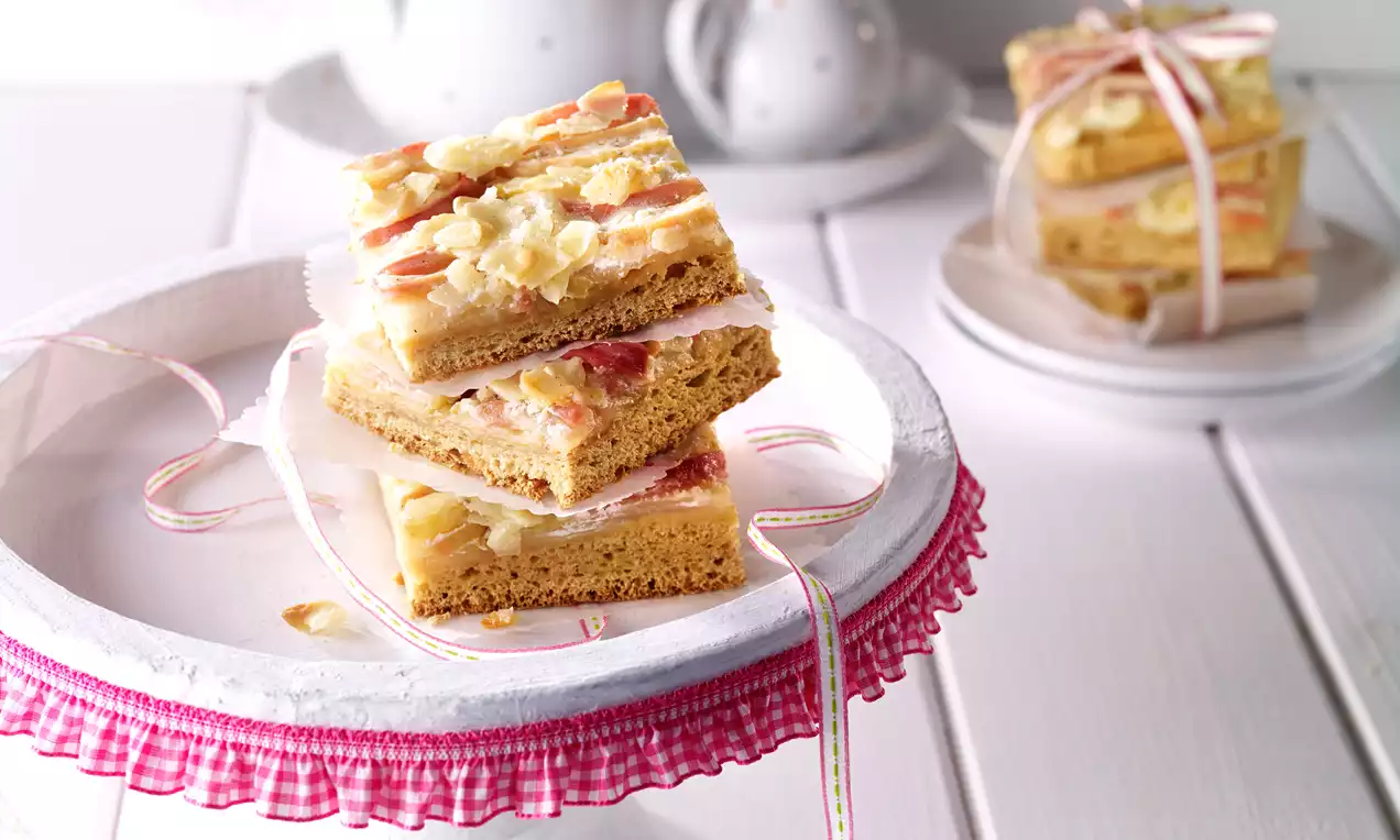 Téglalap formájú, lédús gyümölcsdarabokkal és roppanós mandulaszeletekkel borított sütemény, rétegezve és szalaggal átkötve világos tortaállványon