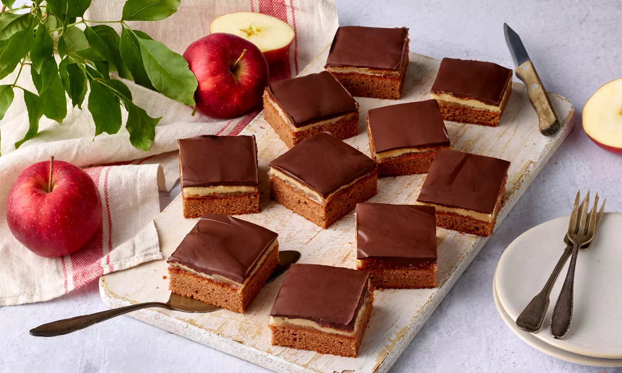 Quadratische Dominostein-Kuchenstücke mit glatter Schokoladenglasur und heller Füllung auf Holzbrett mit roten Äpfeln und Besteck