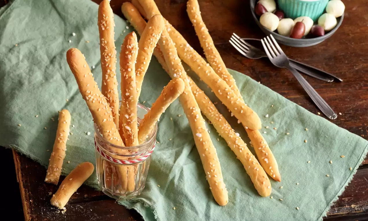 Grissini knusprige goldbraune dünne Stangen, teils mit Sesam bestreut, in einem Glas und gestapelt auf einem Holzbrett neben Schinken und Dip