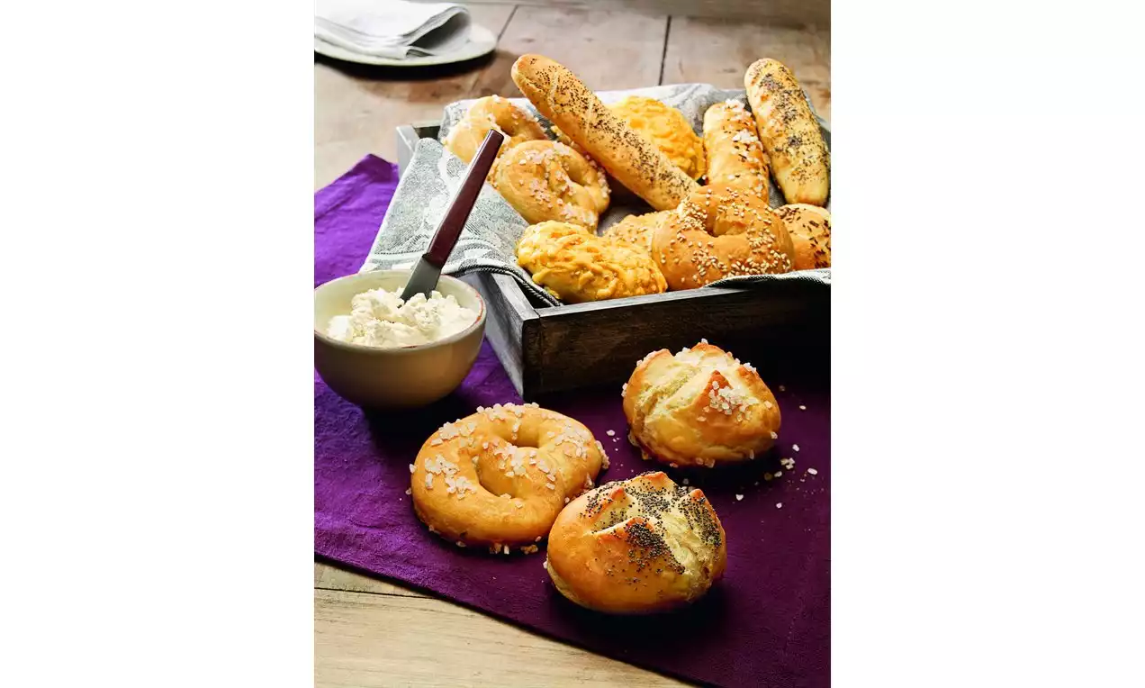 Slana peciva zlaćane boje s hrskavom korom, posuta sjemenkama, raspoređena u drvenu košaru uz zdjelicu svježeg sira na ljubičastom platnu