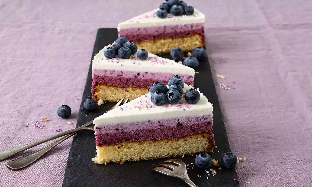 Tranches de gâteau dégradé avec couches violette et crème blanche, myrtilles fraîches en garniture sur plat noir avec fourchettes